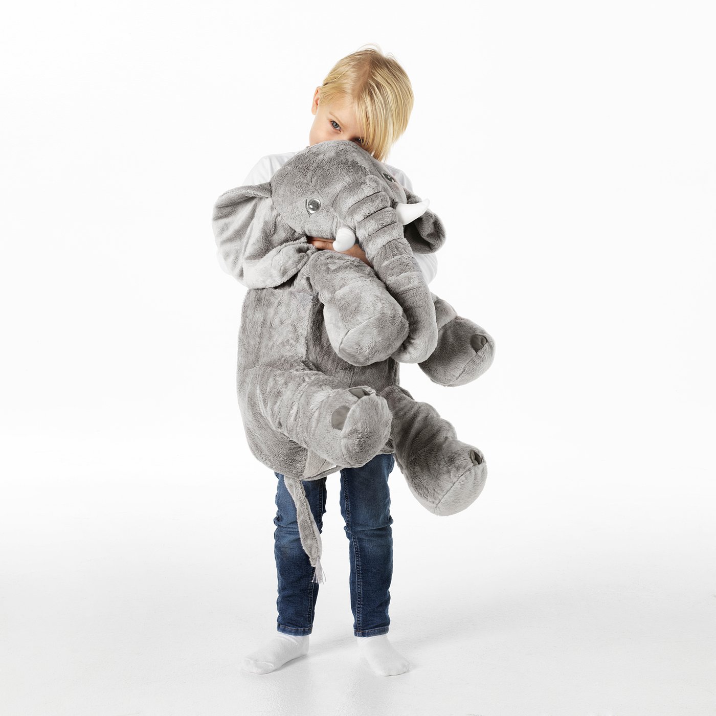 A child hugging a large stuffed elephant toy. The toy is soft, with floppy ears, white tusks, and a cuddly trunk.