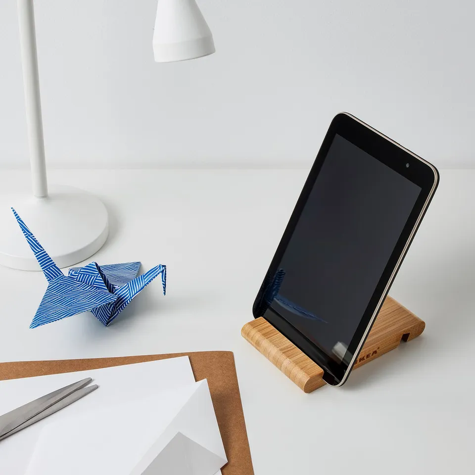 Tablet stand on desk, holding tablet upright. Nearby, a paper crane, lamp, papers, and pen.