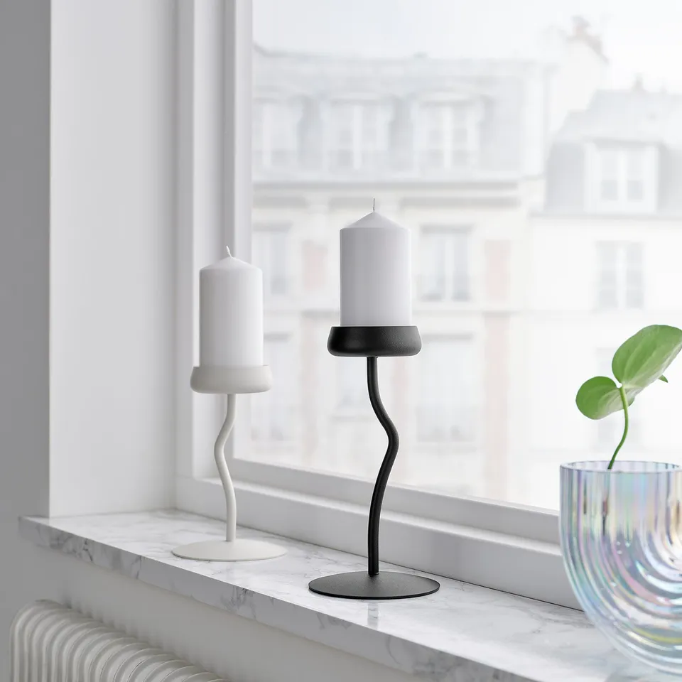 Wavy black candlestick, resembling flower stem, stands tall among white and colourful decor on marble windowsill, next to lime plant.