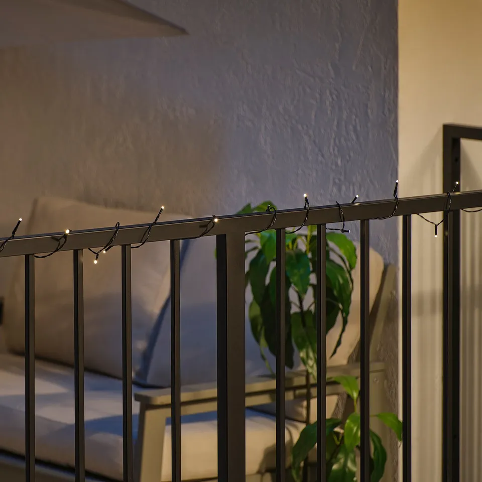 Balcony railing with warm led string lights, creating cosy atmosphere. Plant and outdoor furniture visible.