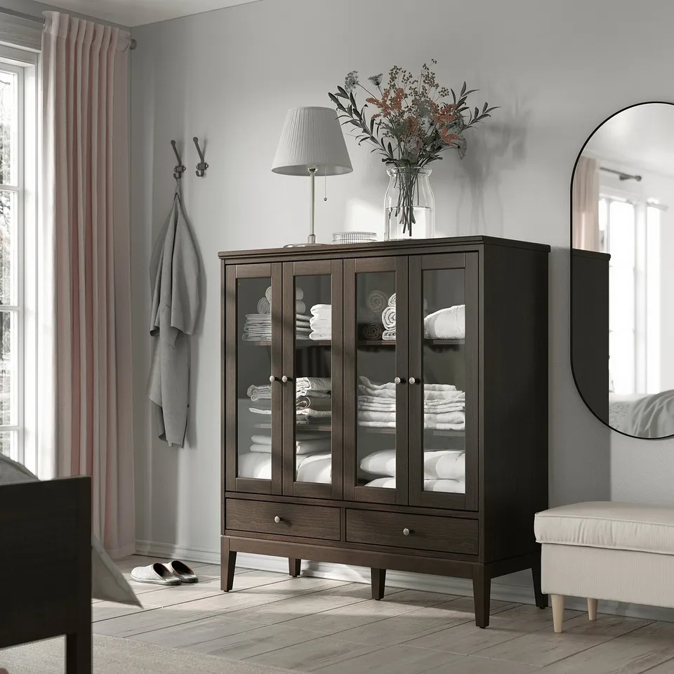 Modern brown wooden cabinet with glass doors, storing folded towels. Topped with a lamp and flowers, beside a mirror and coat hooks.
