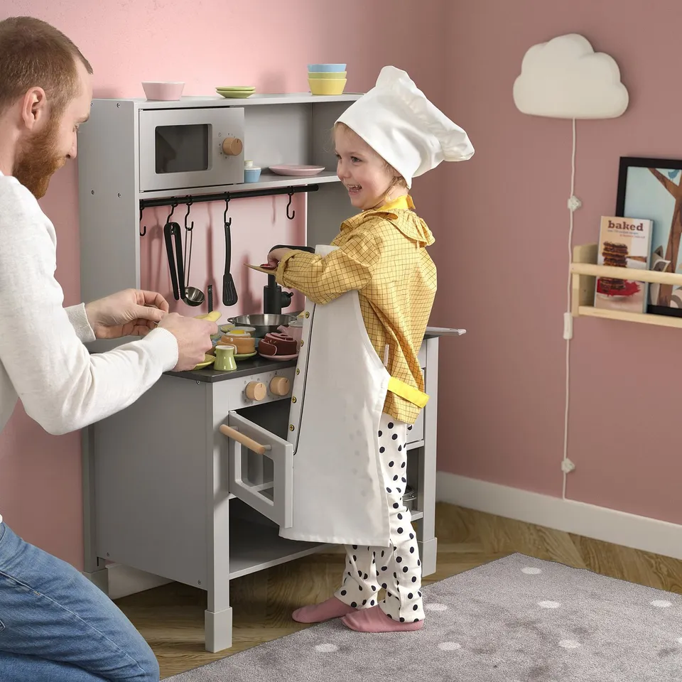 Child cooks in toy kitchen with dad, wearing chef hat and apron, using stove with red lights.