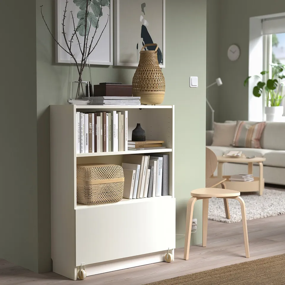 A white BILLY bookcase with a drawer, filled with books and decorated with vases, next to a light wooden stool and sofa in a cosy living room.