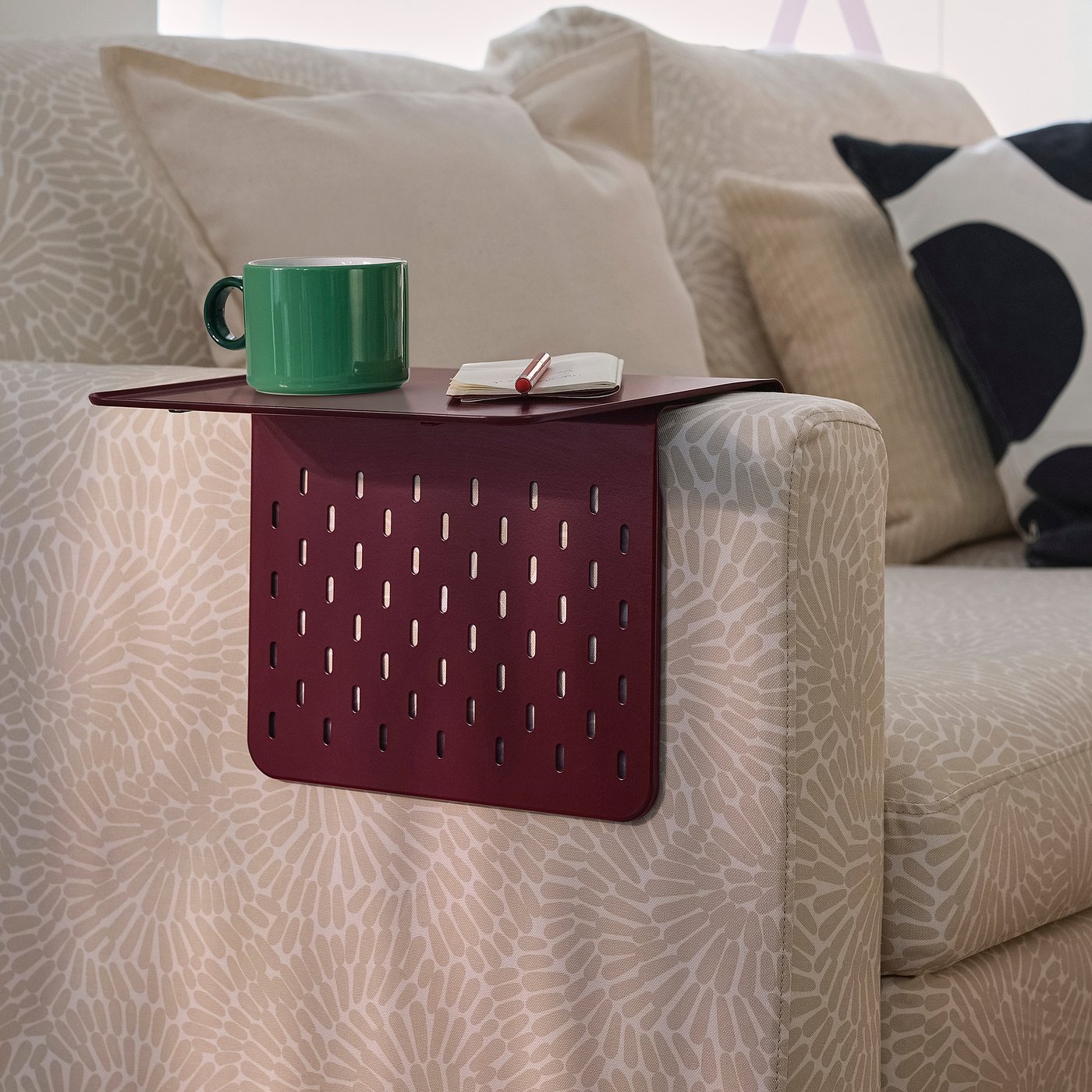 A maroon KRAMPHULT armrest tray on a sofa, holding a green mug and a book, offering a convenient spot for relaxation.