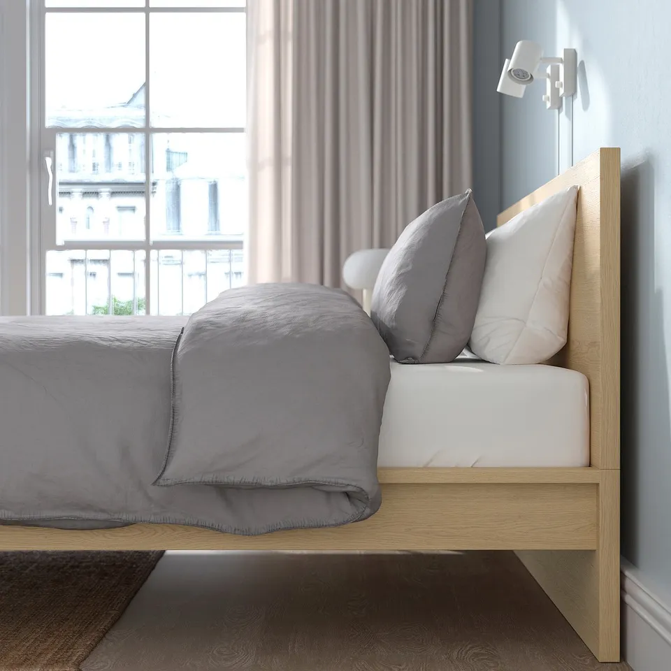 Bedroom with MALM bed featuring high headboard, slatted base, and adjustable bed sides. Grey bedding, pillows, window curtain, and wall sconce.