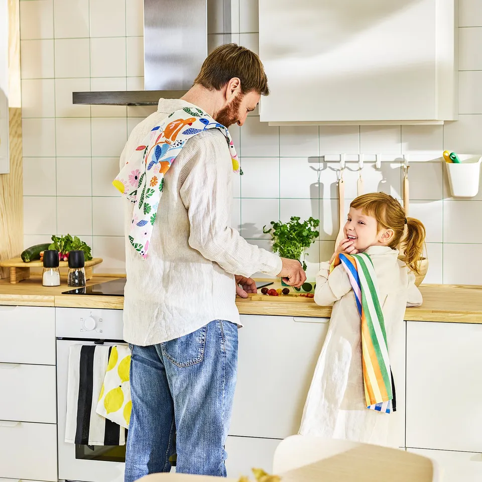 Happy kitchen scene: person and child in aprons, laughing. Bright towels hang in the well-lit kitchen.