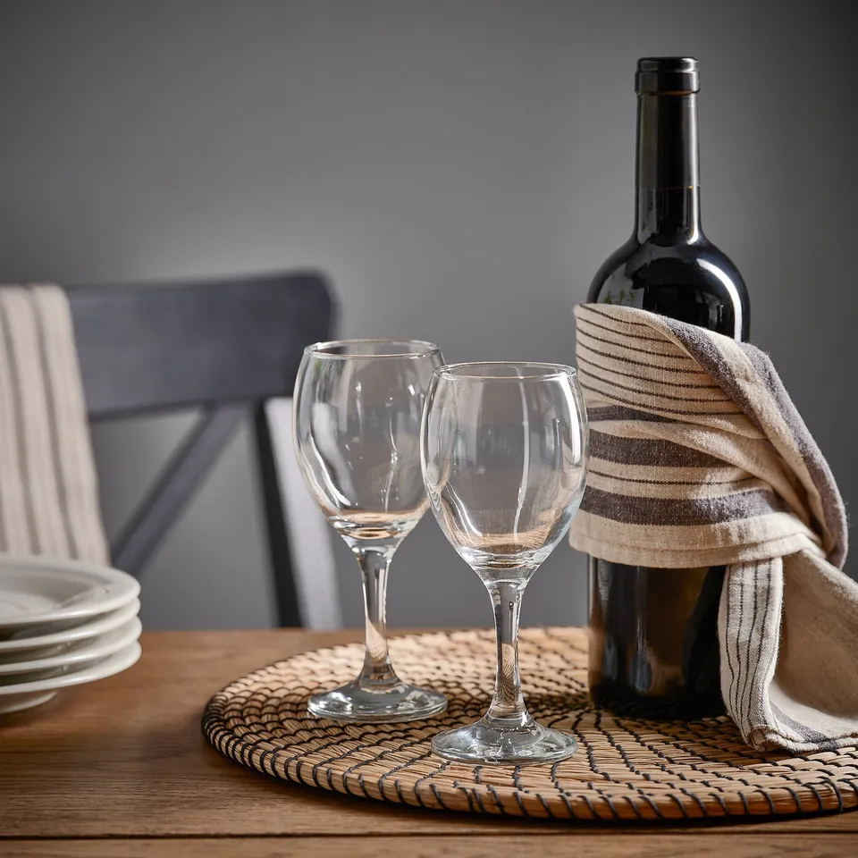 Two ÄNKEBLOMSTER wine glasses, a bottle, and a cloth on a woven mat, set on a wooden table.