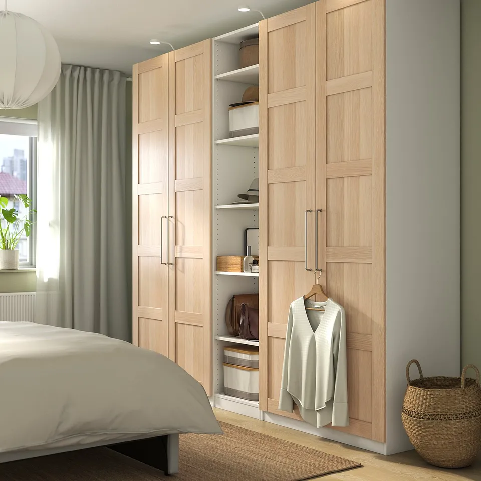 Wooden PAX wardrobe with open door showing clothes. Beside bed, near window and basket.
