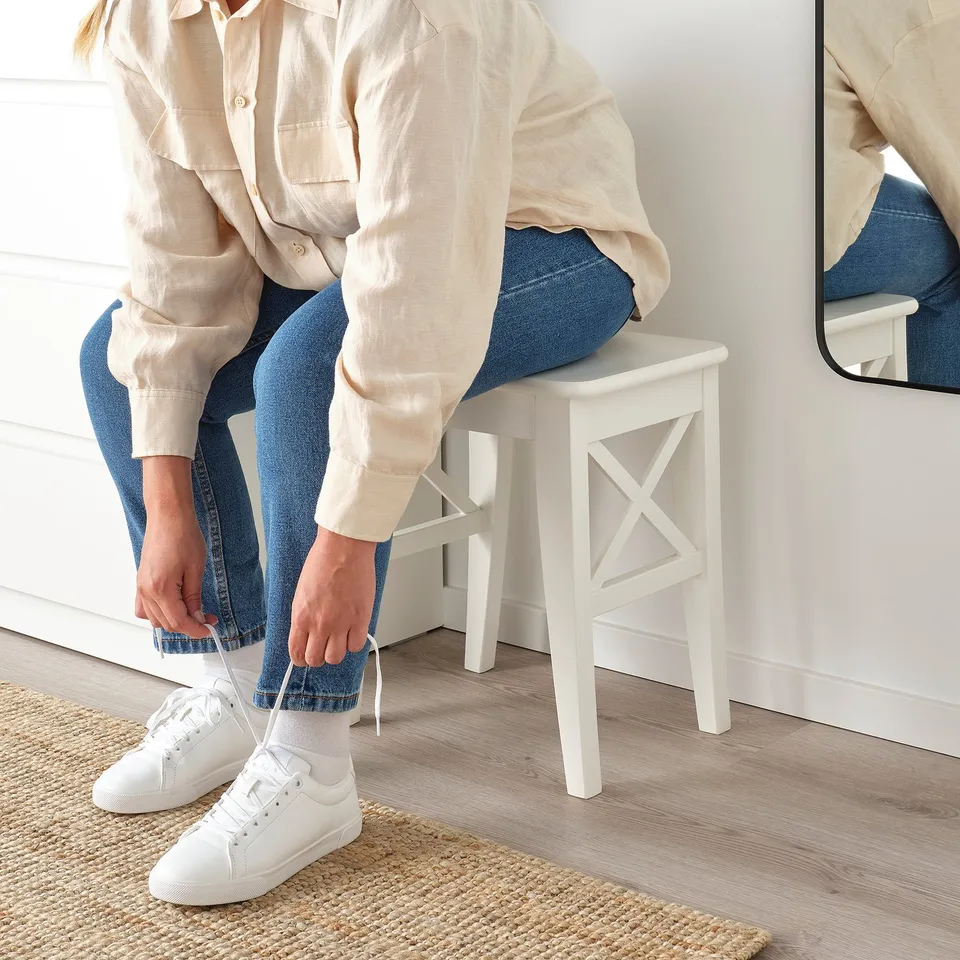 Person tying white shoelaces, seated on white ROSENTORP stool, wearing light shirt, blue jeans, and white trainers. Natural-fiber rug beneath.