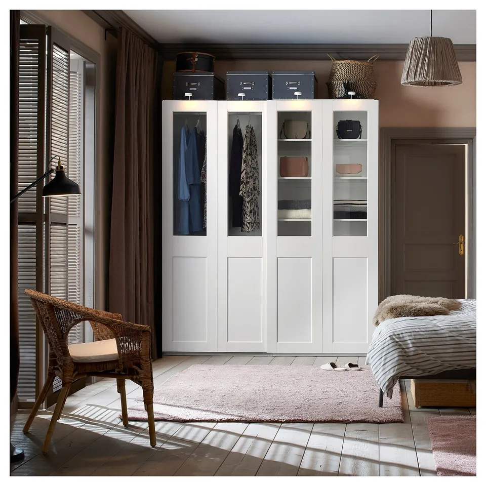 Modern bedroom with white PAX wardrobe, wicker chair, and rug. Clothes displayed behind glass doors.