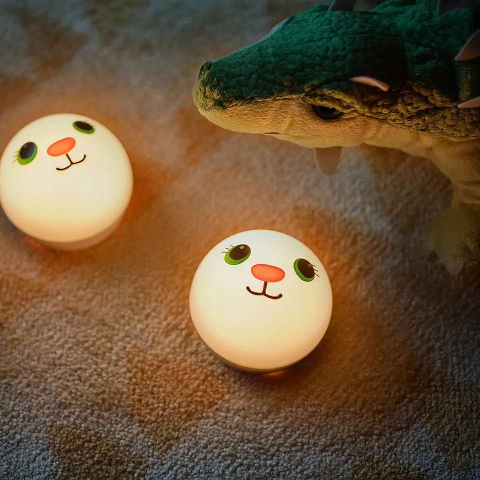 Two small white spherical nightlights with cartoon faces, green eyes, and orange noses sit on a carpet, next to a green dinosaur toy.