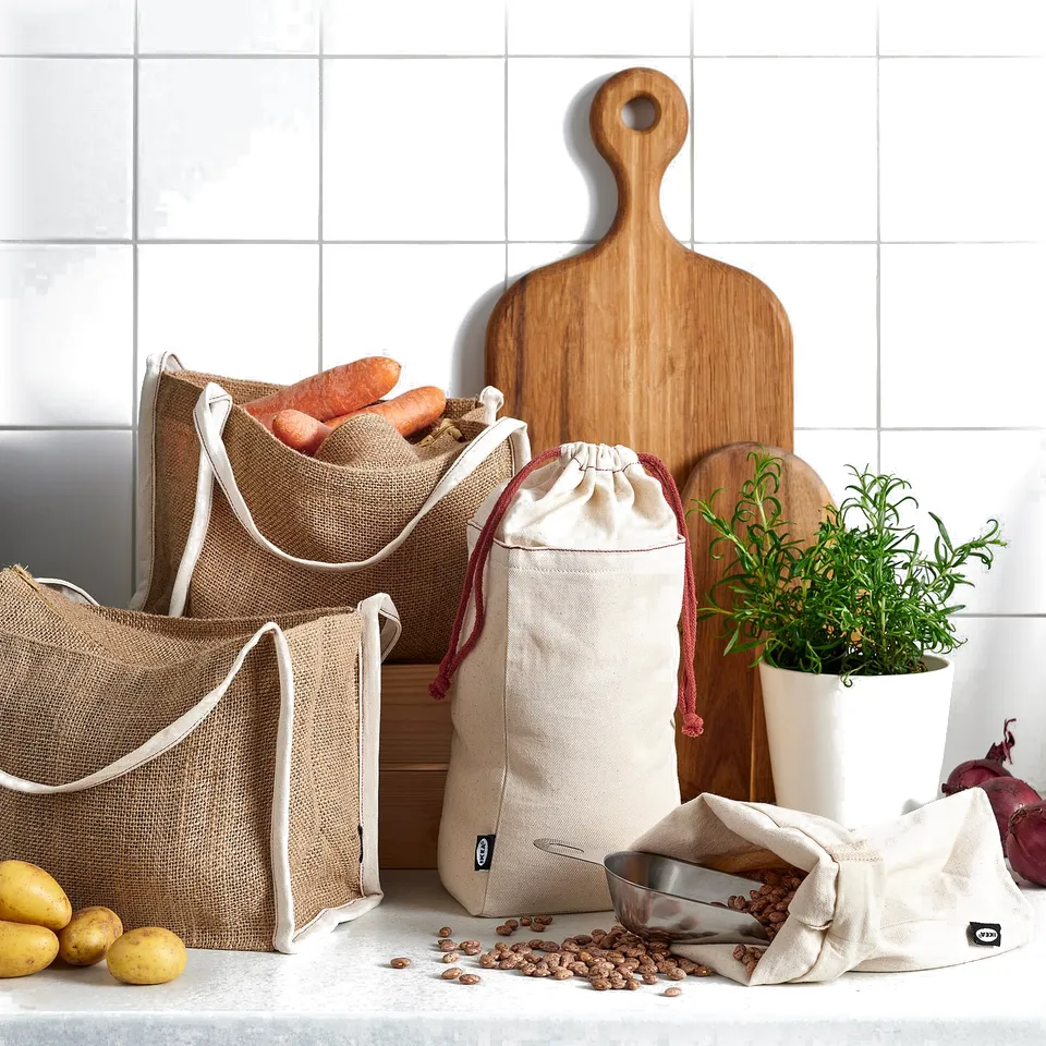 Natural hessian bags with white trim store root vegetables, beans. Wooden chopping boards, potted herb, red onion nearby.
