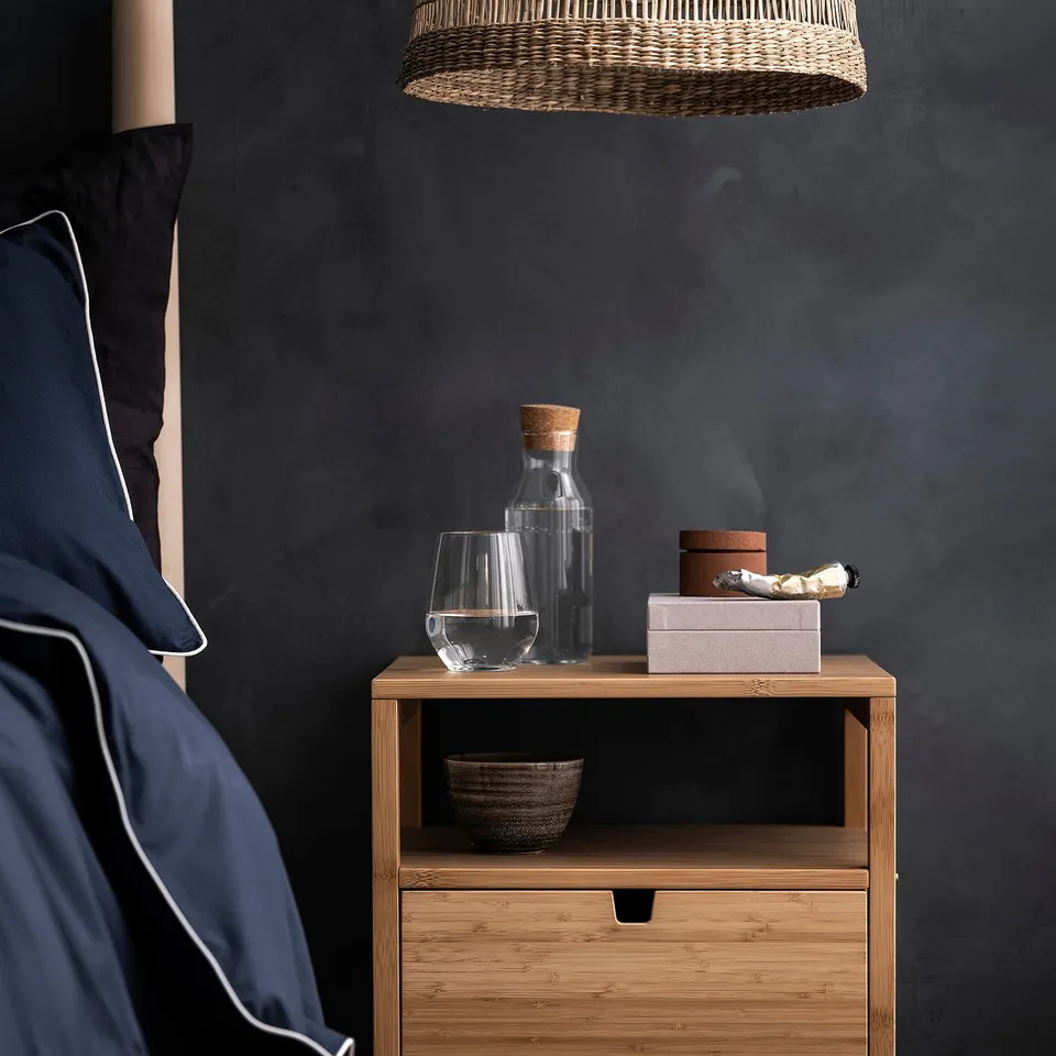 Small glass carafe with cork lid on wooden bedside table, next to a glass cup and other items, against a dark wall with a wicker light pendant.