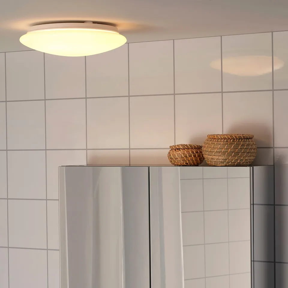 Modern bathroom with a circular ceiling light providing even illumination. Two wicker baskets sit on a glossy cabinet under light tiles.