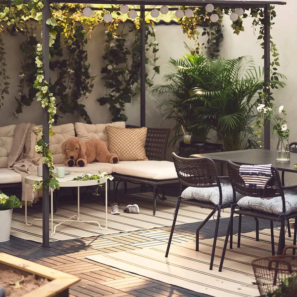 Cosy outdoor patio with beige GUNNÖN pergola, wicker furniture, cushions, blankets, potted plants, and string lights.