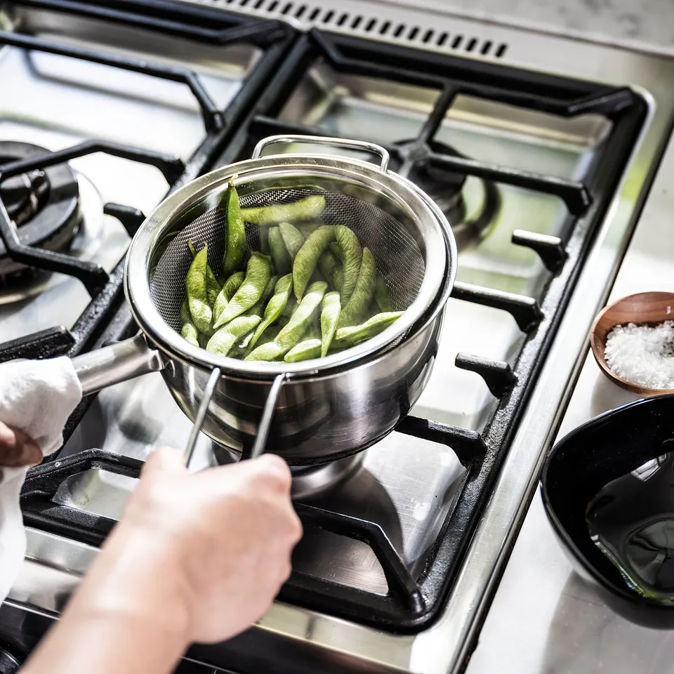 A person holds a silver metal strainer full of green vegetables over a gas stove.
