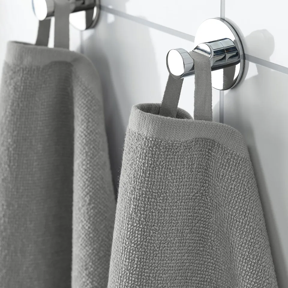 Two grey towels hang from a chrome hook on a white wall. The towel in front is folded neatly, showing its texture and lightweight fabric.