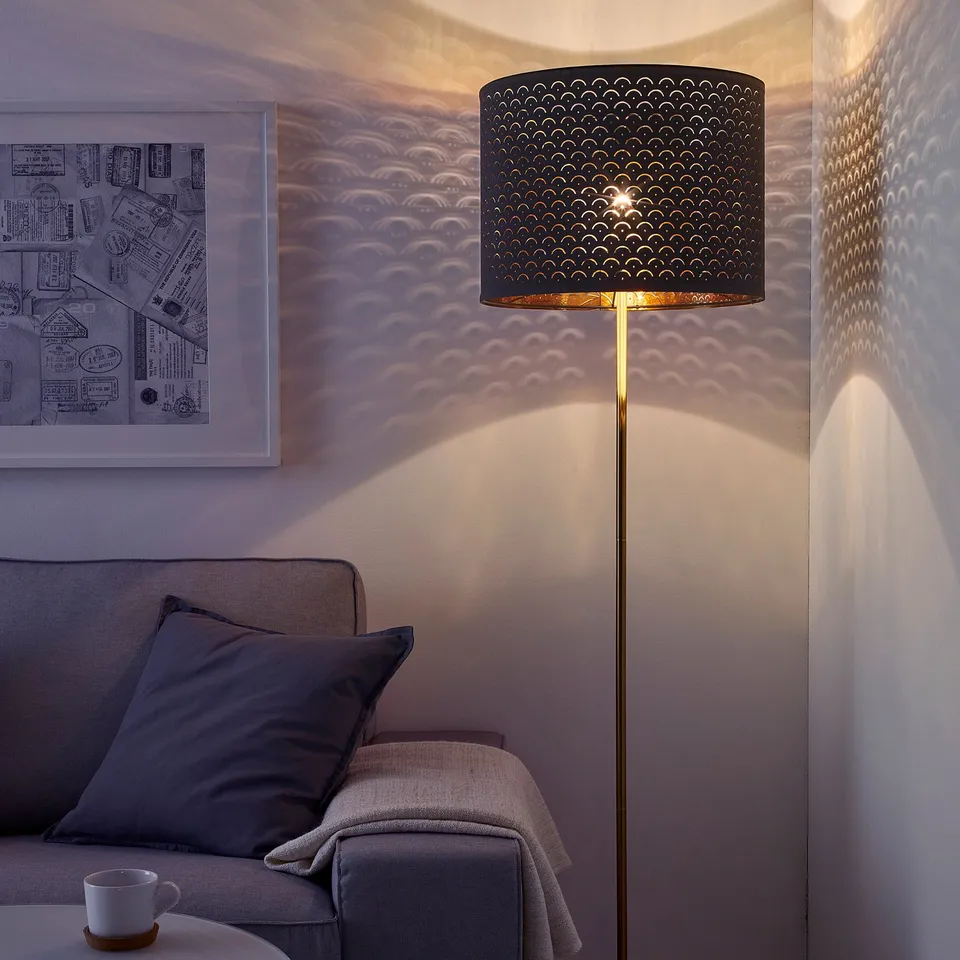 A cosy living room with a lit floor lamp casting intricate patterns on the wall. A sofa is nearby with a mug on a side table.