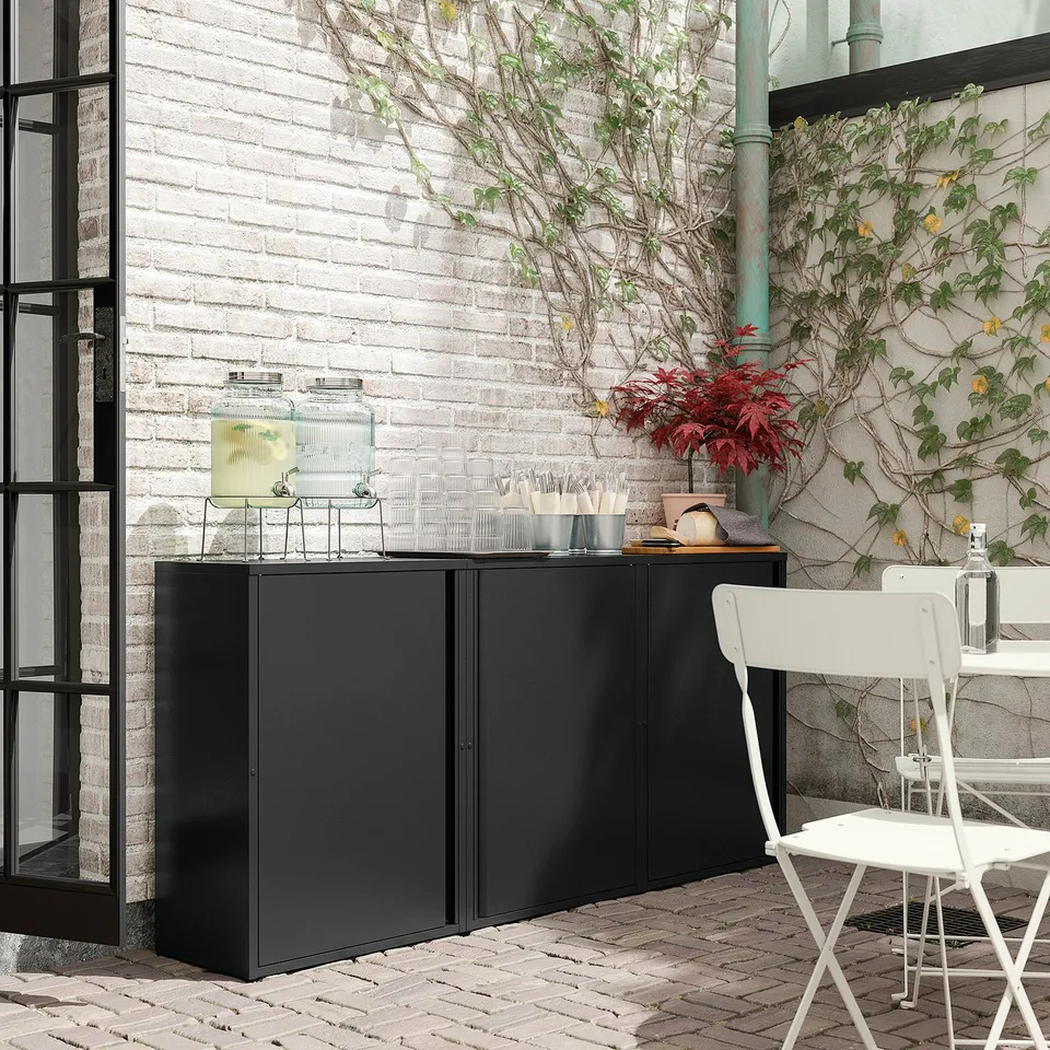 Black metal cabinet with 2 doors & shelves, adjustable feet, outdoors against white bricks, with chair & jars.