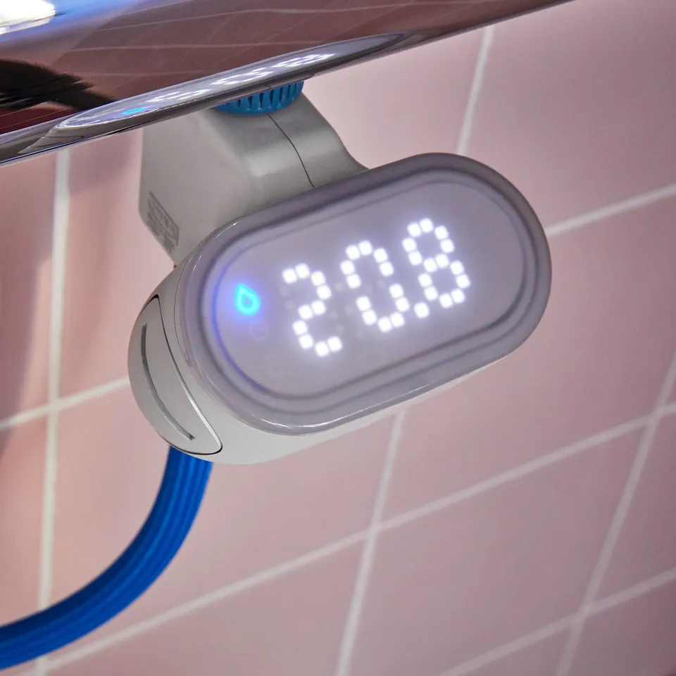 Digital water metre showing 3:00, attached to blue hose, mounted on pink tiled wall. Displays water usage and temperature.