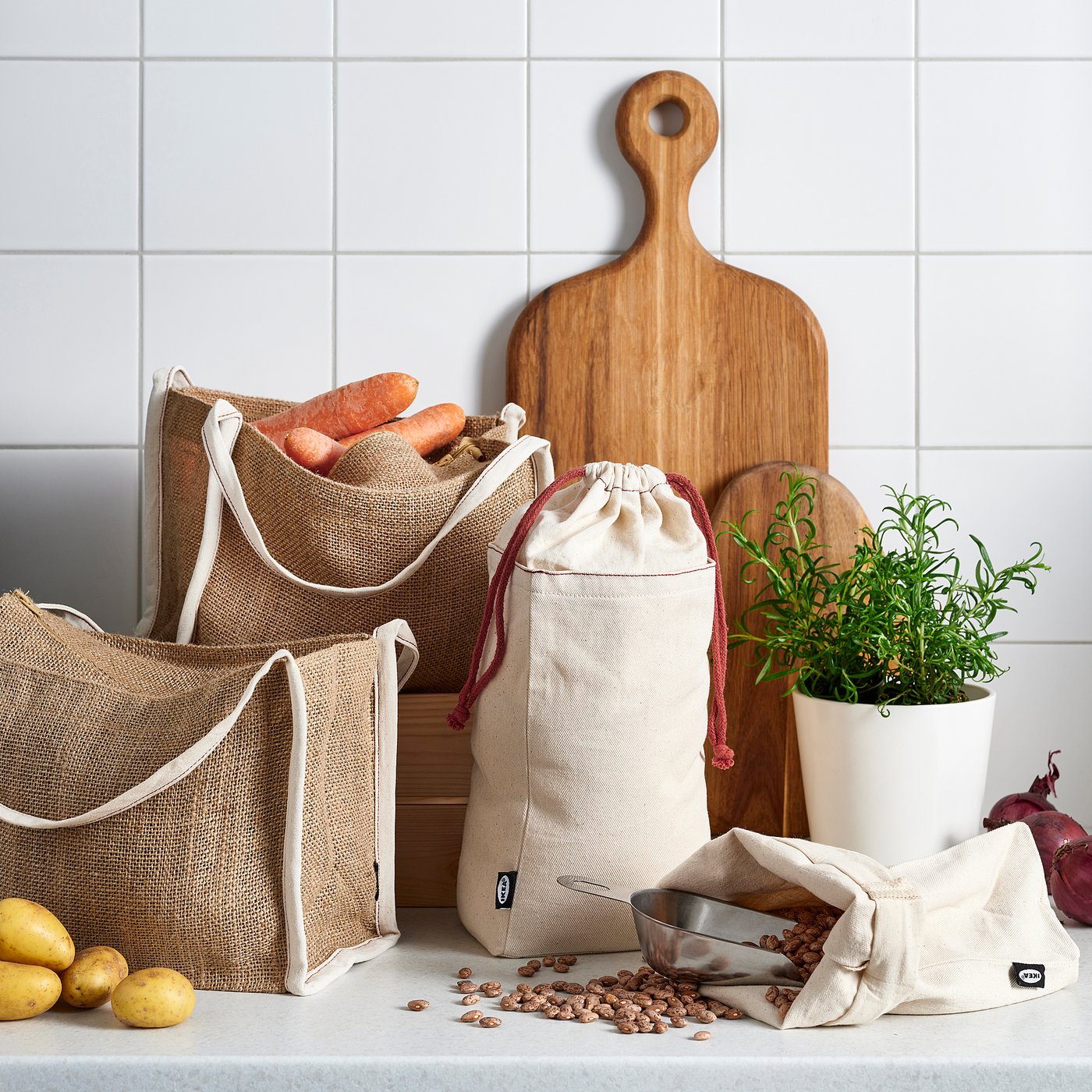 Beige jute bags with white handles store veggies, cutting boards, and herbs on kitchen counter.