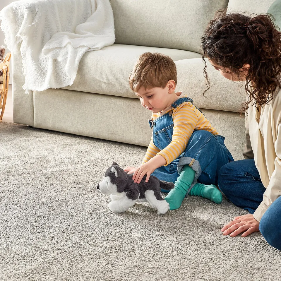 Child playing with soft toy husky on carpet, adult nearby. Toy has bright blue eyes, safe for ages 12 months +.