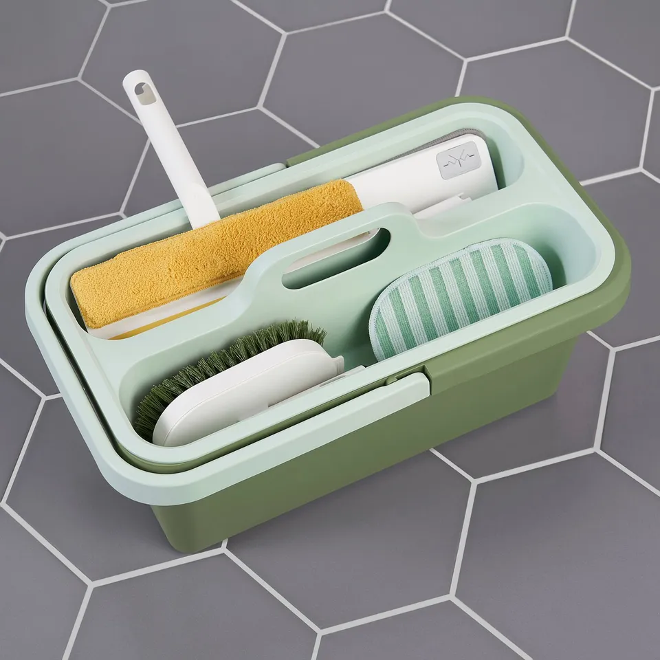 Green cleaning bucket with handle, storing tools like a brush, sponge, and mop, has a textured base for washing.