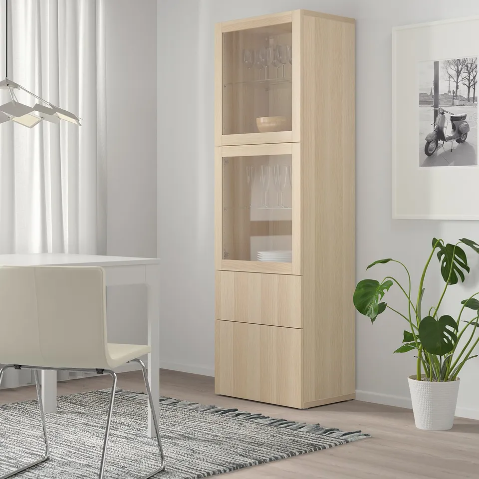 Tall BESTÅ cabinet with wine glasses & plates, next to table & chairs on rug, near plant & photo.