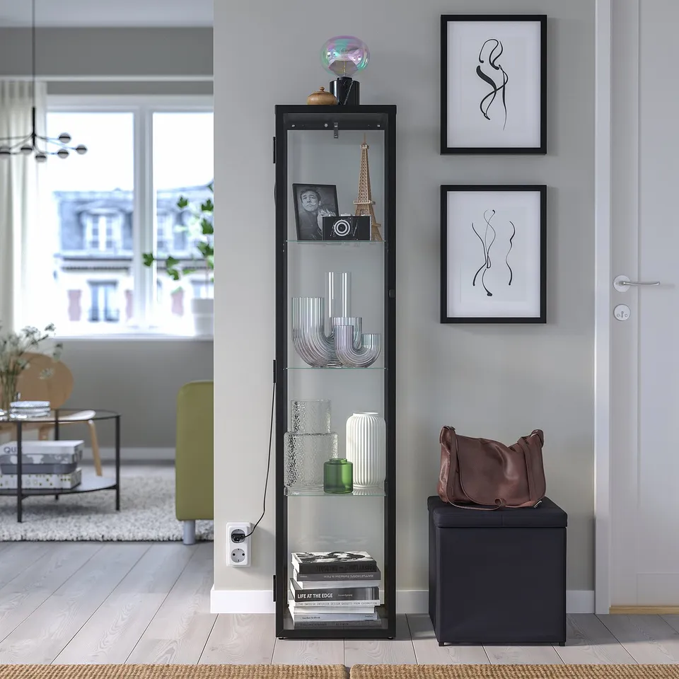 BLÅLIDEN glass cabinet with shelves, holding vases, camera, and pictures. Beside doorway, contemporary furniture, cushioned seat, and bag.