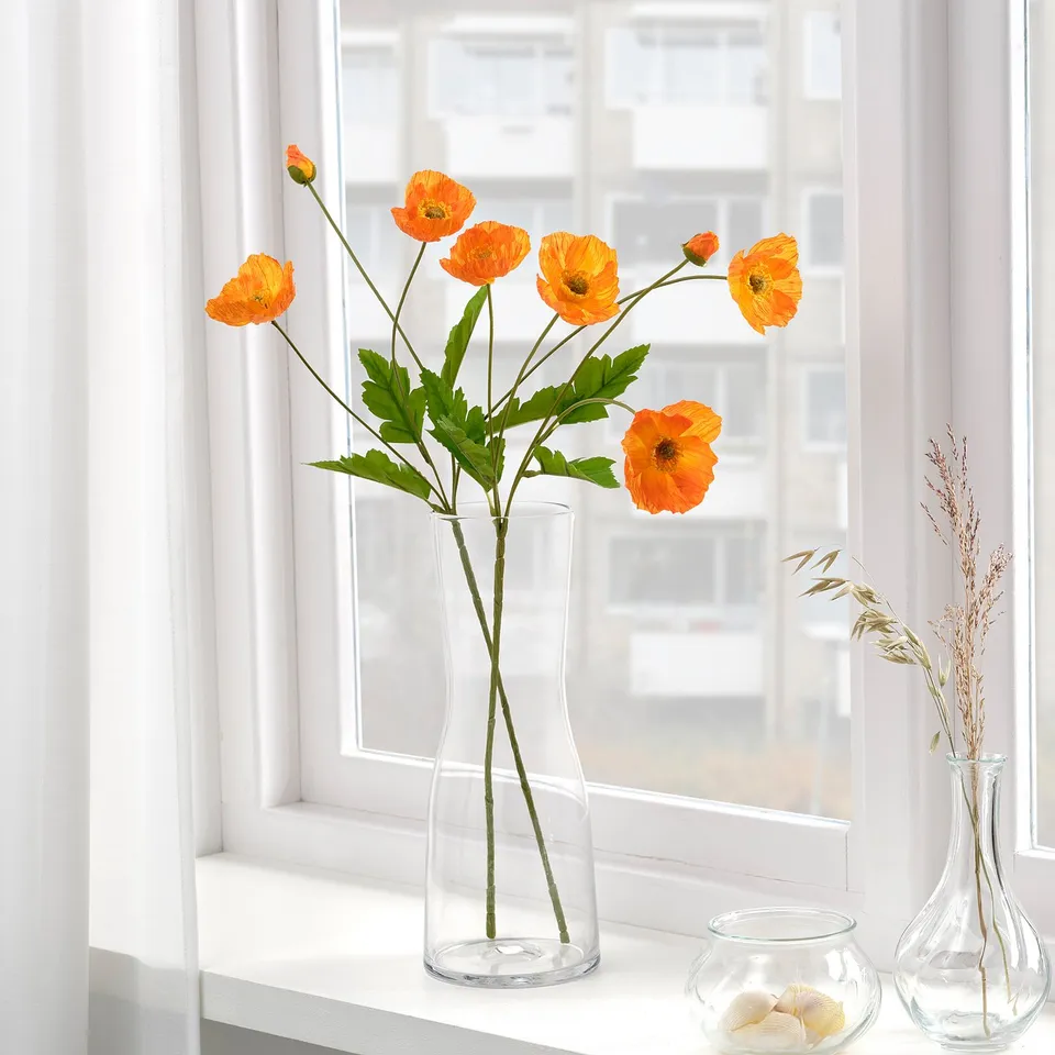 Bright orange artificial flowers in a clear glass vase sit on a windowsill, adding vibrant colour to the scene.
