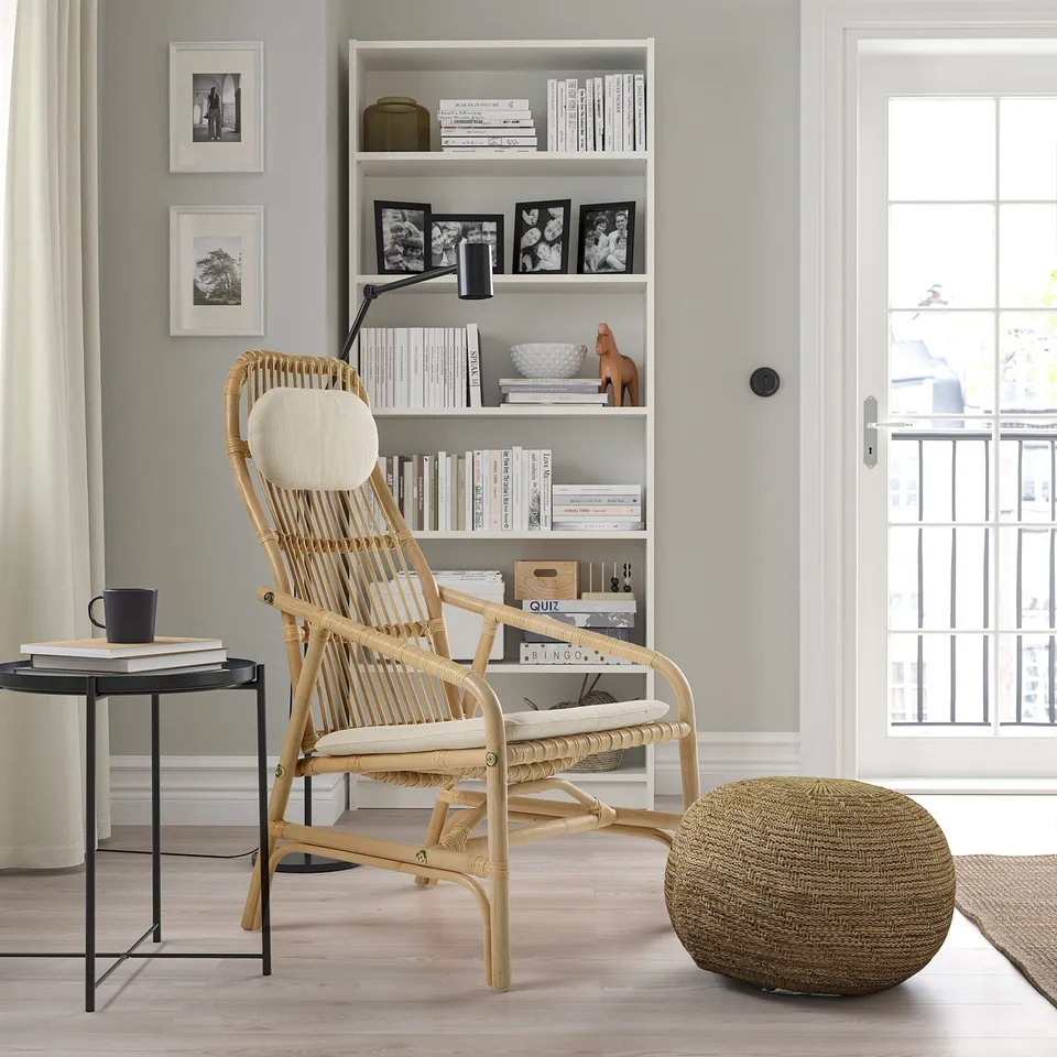 Cosy nook with wicker armchair, cream cushion, side table, books, mug, tall bookshelf, window, round ottoman.