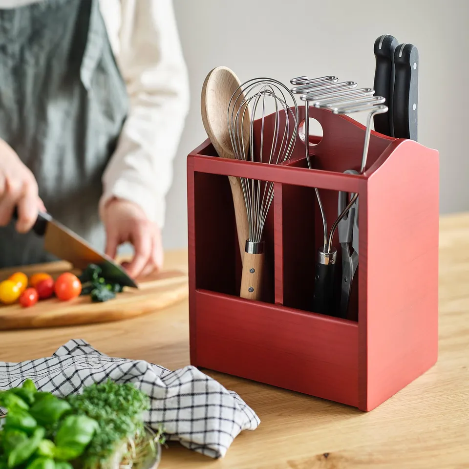 Red wooden knife block holding kitchen utensils and knives, next to cutting board and fresh herbs on table.