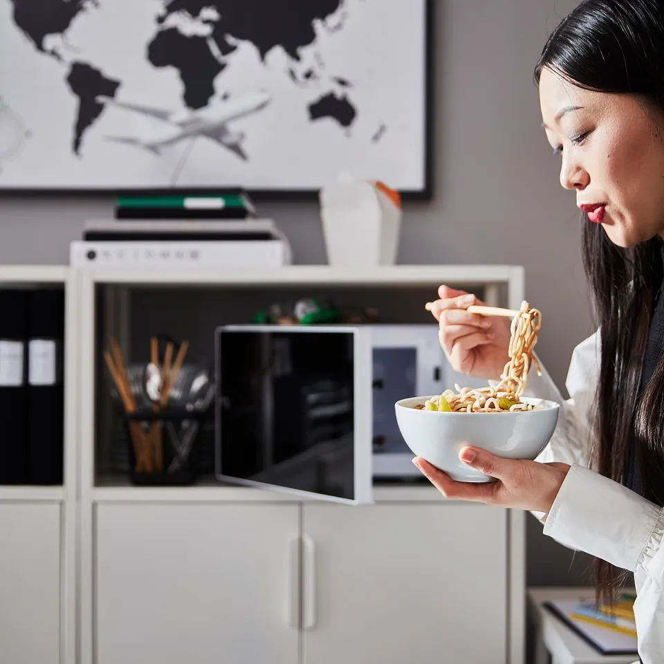 A person eats noodles from a bowl near a white TROTTEN storage unit, which has shelves and drawers, holding items like chopsticks and a microwave.
