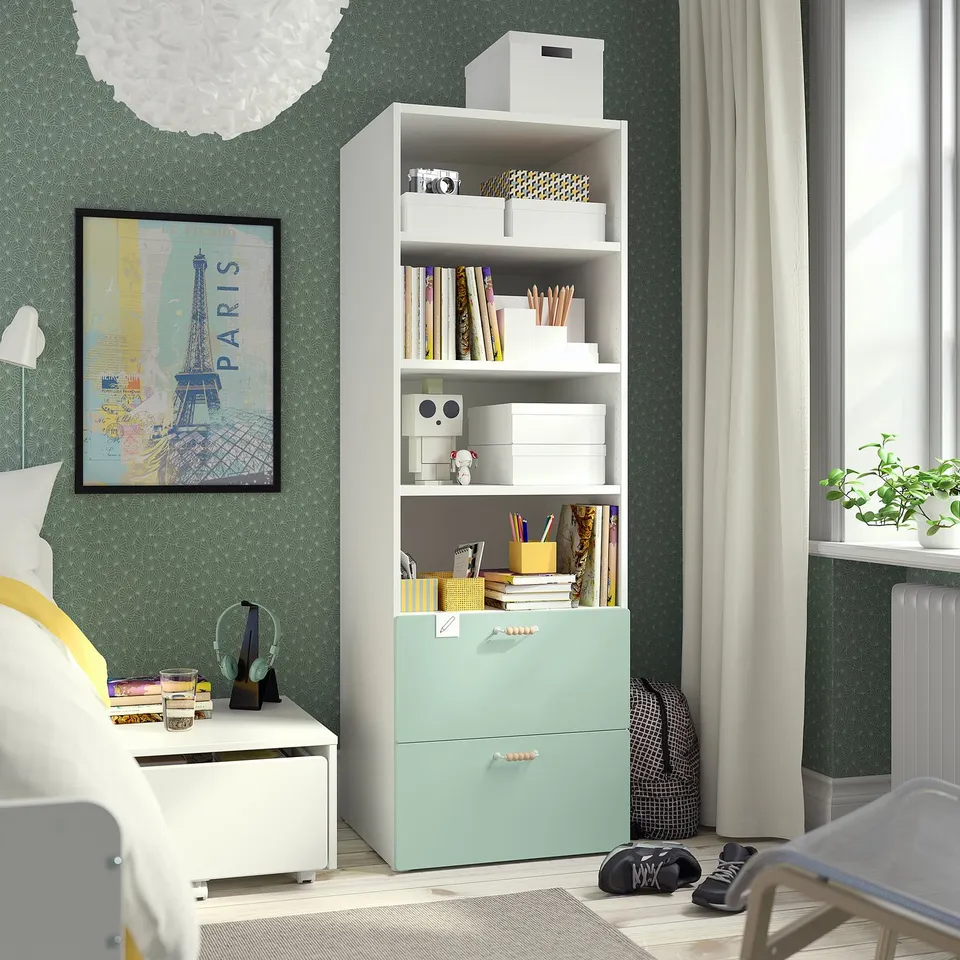 A SMÅSTAD bookcase in mint green with various storage items. Bright and compact, it fits well next to a bed with a map poster behind.