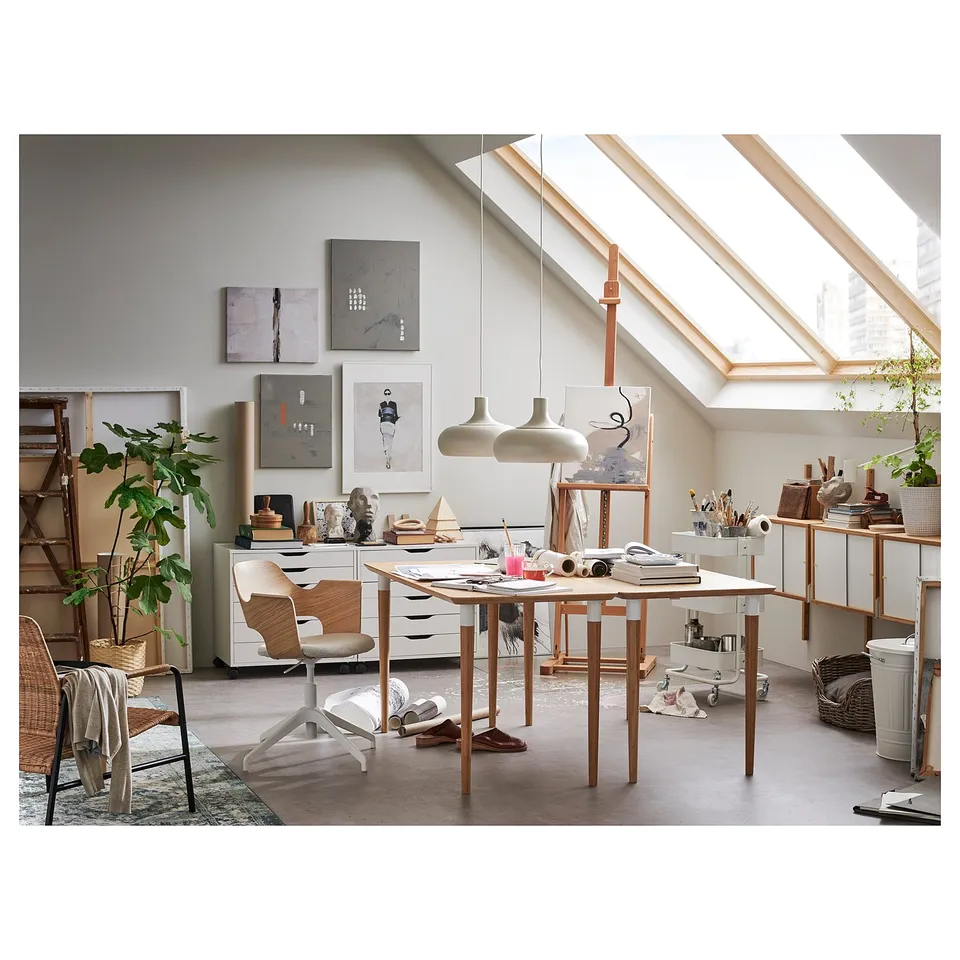 Art studio with slanted ceiling, large windows, wooden desk, chair, shelves, art supplies, and paintings.