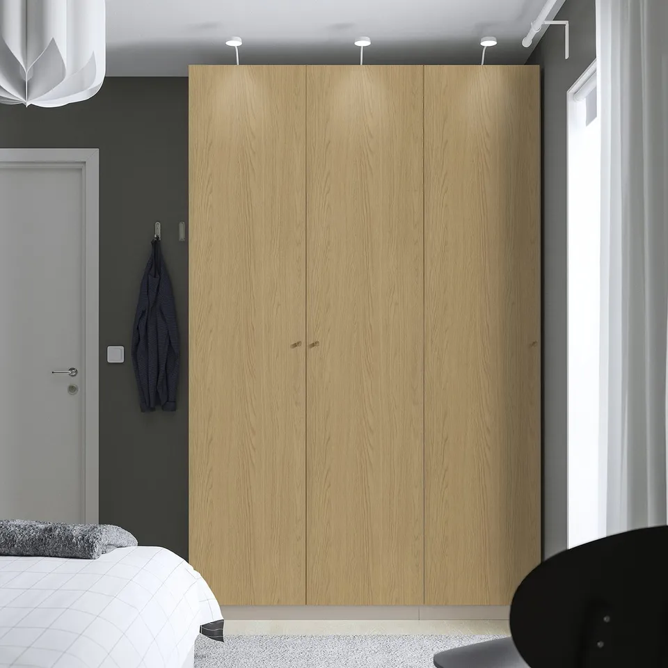 Modern minimalist bedroom with large wooden closet between white door and window, hanging blue jacket and white bed.