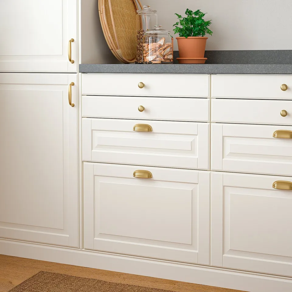 White kitchen cabinets with brass handles; countertop holds jars and a potted plant.
