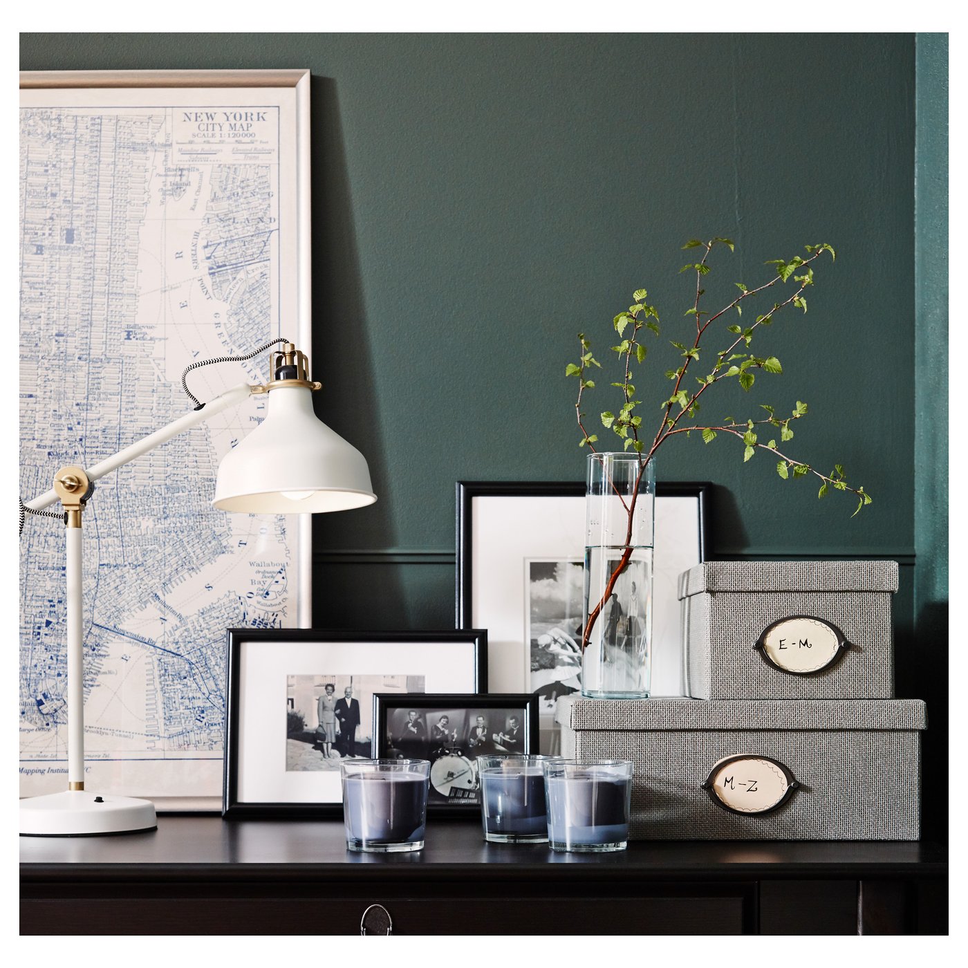 Desk setup with adjustable lamp, photo frames, boxes, and a tall clear vase holding green branches.