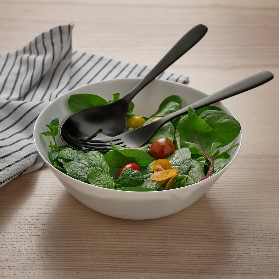 Black cutlery in white bowl with salad on wooden table.