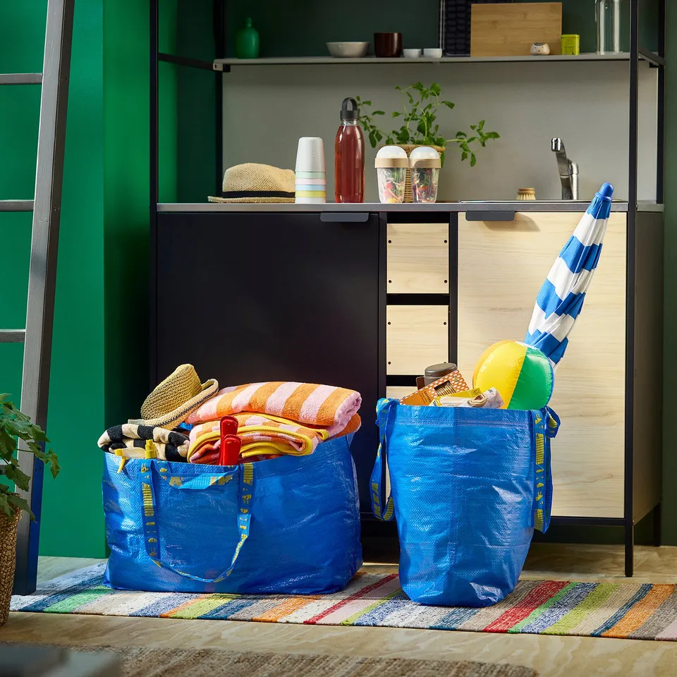 Two blue IKEA FRAKTA bags filled with beach towels, toys, and an umbrella on a colourful rug near a green shelf with kitchen items.