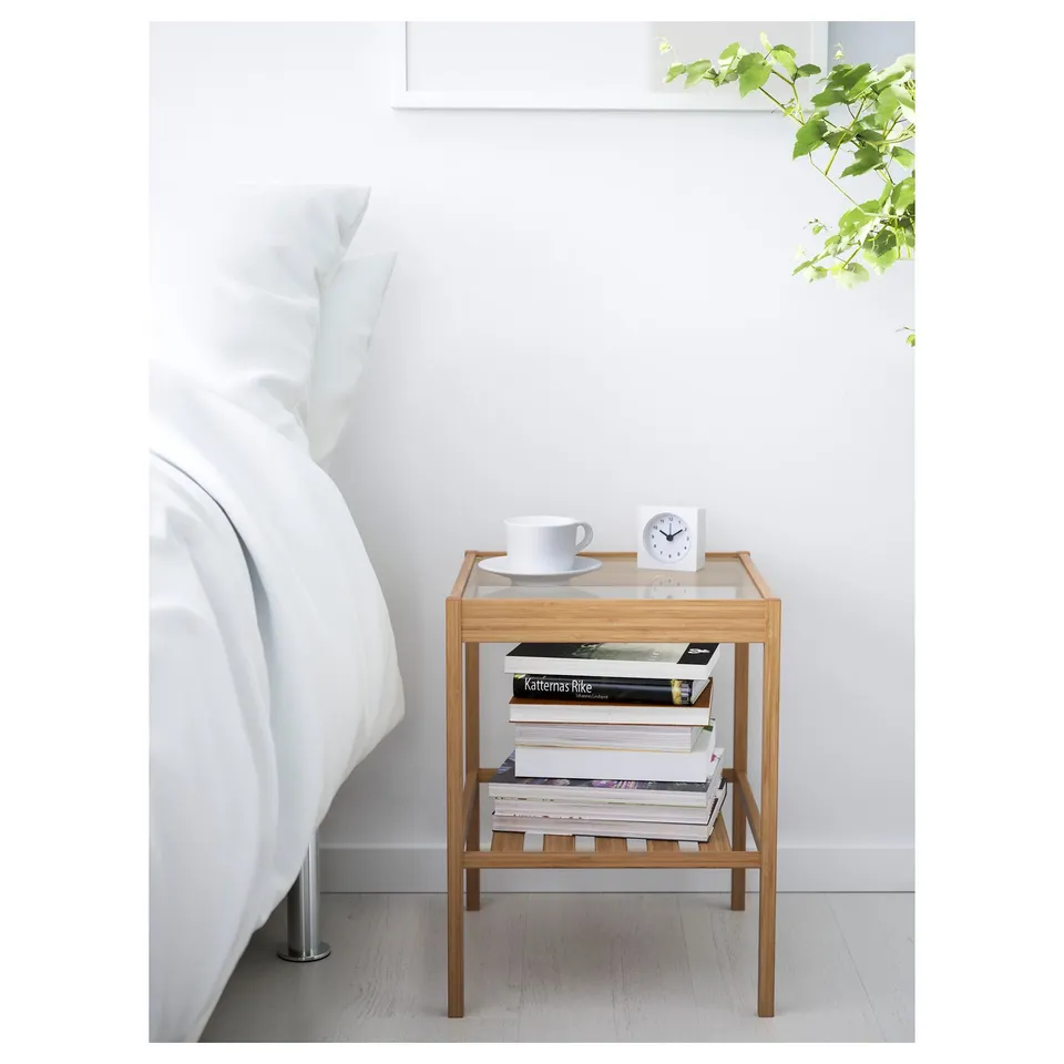 Small wooden bedside table with glass top, holding books, a cup, and alarm clock, placed next to a white bed.