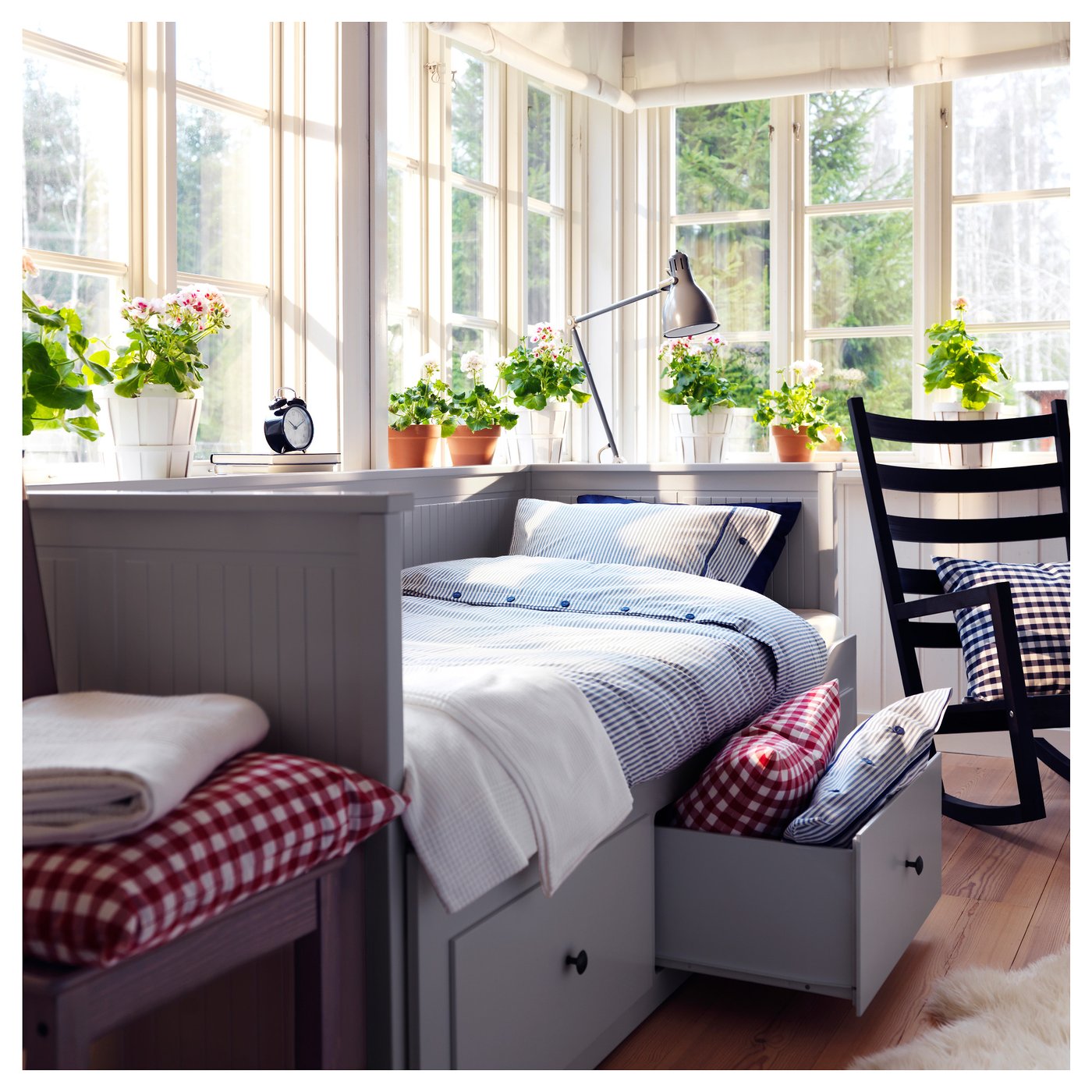 Bright, cosy bedroom with white HEMNES day-bed, plaid pillows, plants, large windows, black rocking chair, and furry rug.
