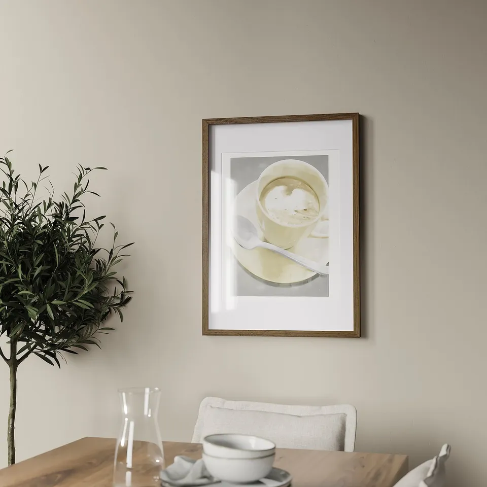 A minimalist living space with a framed picture of coffee. The wooden frame hangs on a wall above a wooden table with a jug, small bowl, and plant.