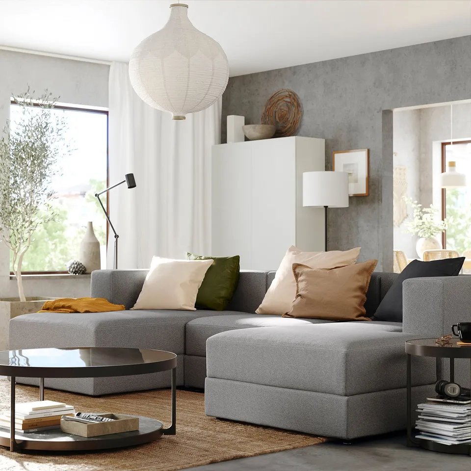 Modern living room with grey JÄTTEBO sectional sofa, colourful pillows, wooden coffee table, and large window with curtains.