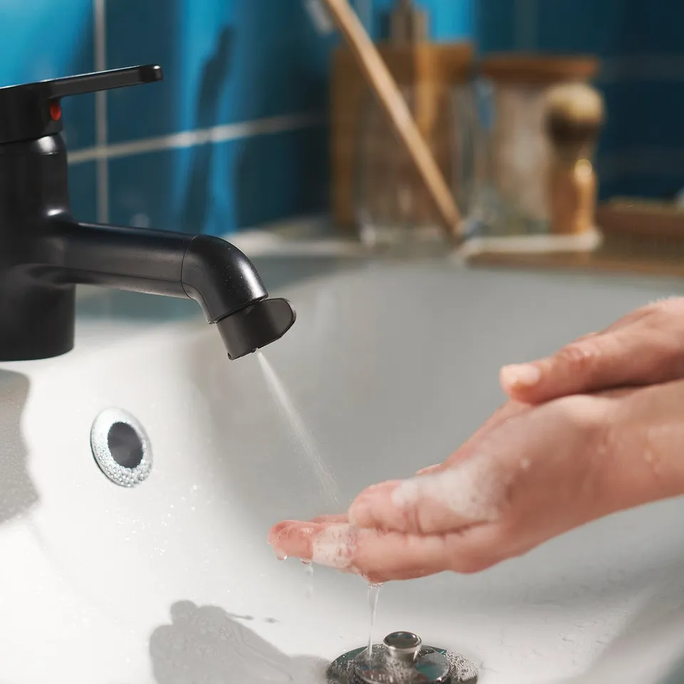 A person washing their hands under a black ikea ÅBÄCKEN sink faucet with a spray nozzle, set against a blue tiled wall.