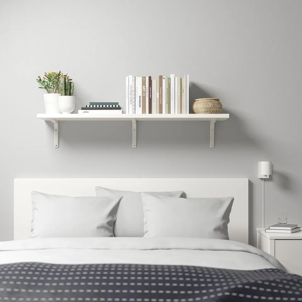 Modern bedroom with white BERGSHULT shelf over bed, holding books, basket, and cactus.