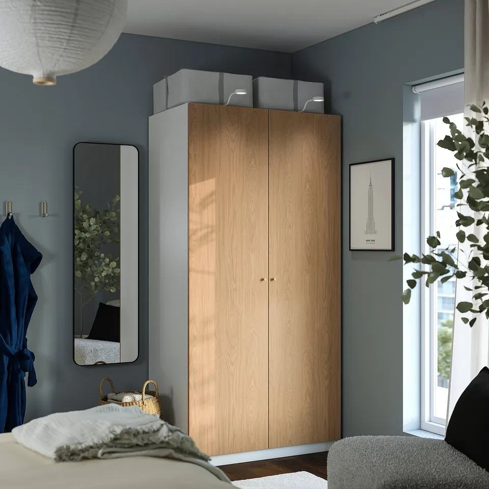 A modern bedroom with a light wooden PAX wardrobe against a dark blue wall, featuring a mirror, window, and hanging plant.