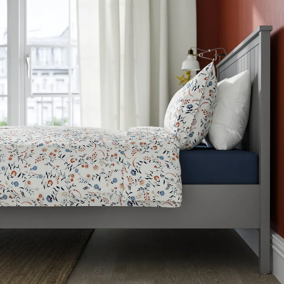 A neatly made bed with a white floral pattern comforter, two pillows, and a grey headboard. Soft lighting illuminates the serene room.