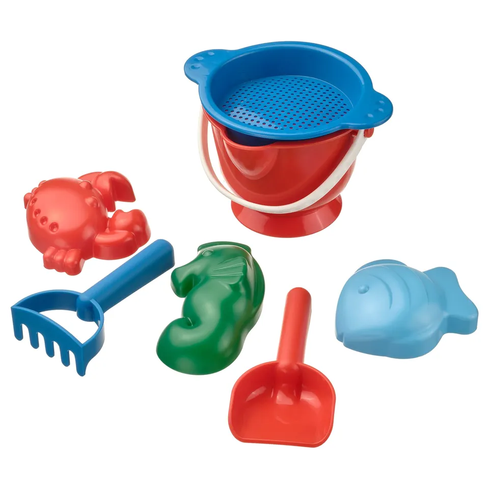 A colourful beach toy set includes a red and blue bucket, crab, rake, green dinosaur, red shovel, and blue fish. Plastic, sand toys, fun design.