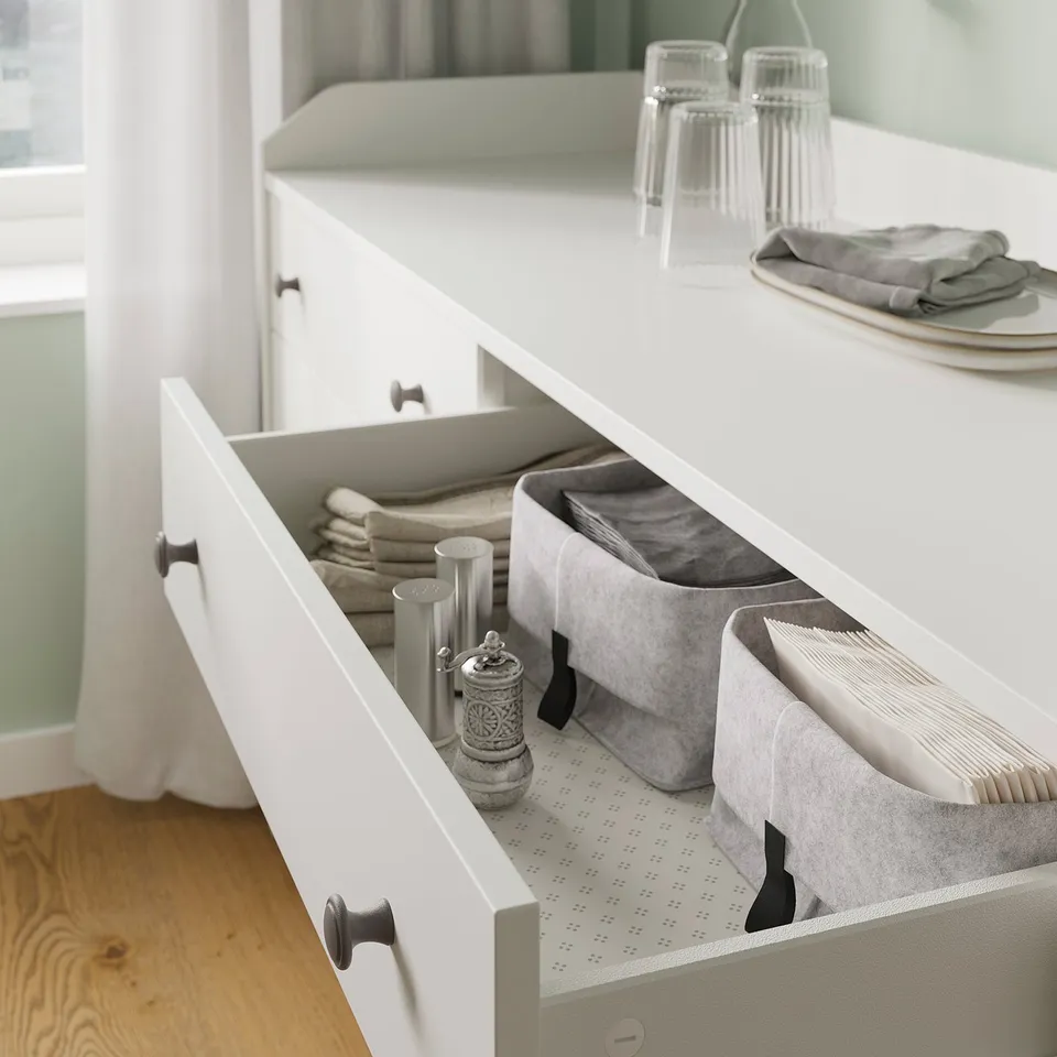 Open white HAUGA drawer, organised with fabric bins, stacked napkins, and seasoning jars, on wooden floor.